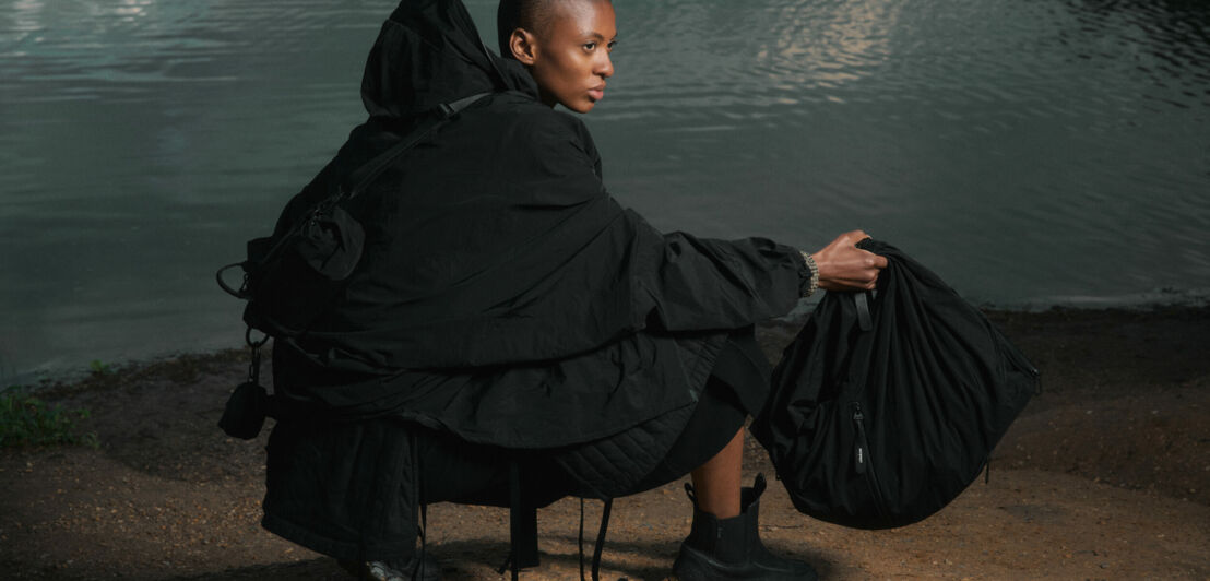 In Schwarz gekleidete Frau mit Taschen von côte&ciel in der Hand und auf dem Rücken vor einer Kulisse am See