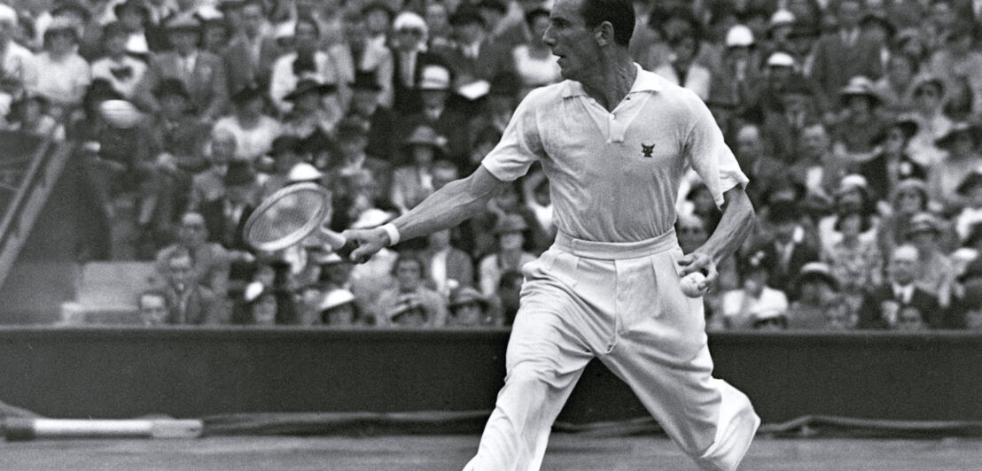 Schwarzweißaufnahme von Fred Perry in weißer Sportkleidung bei einem Tennisspiel vor Publikum