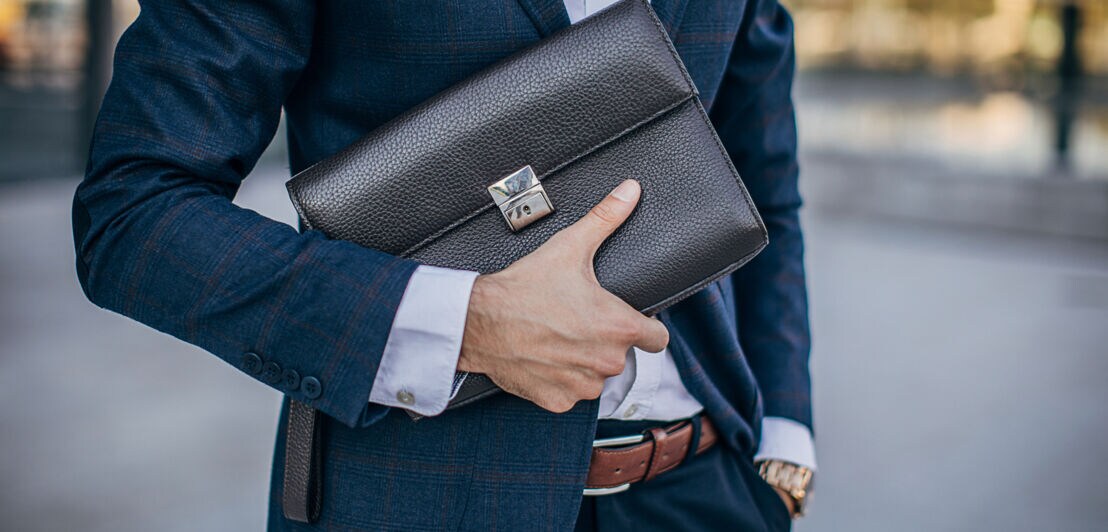 Detailaufnahme einer Person im Anzug, die eine Männerhandtasche aus Leder in der Hand hält. 