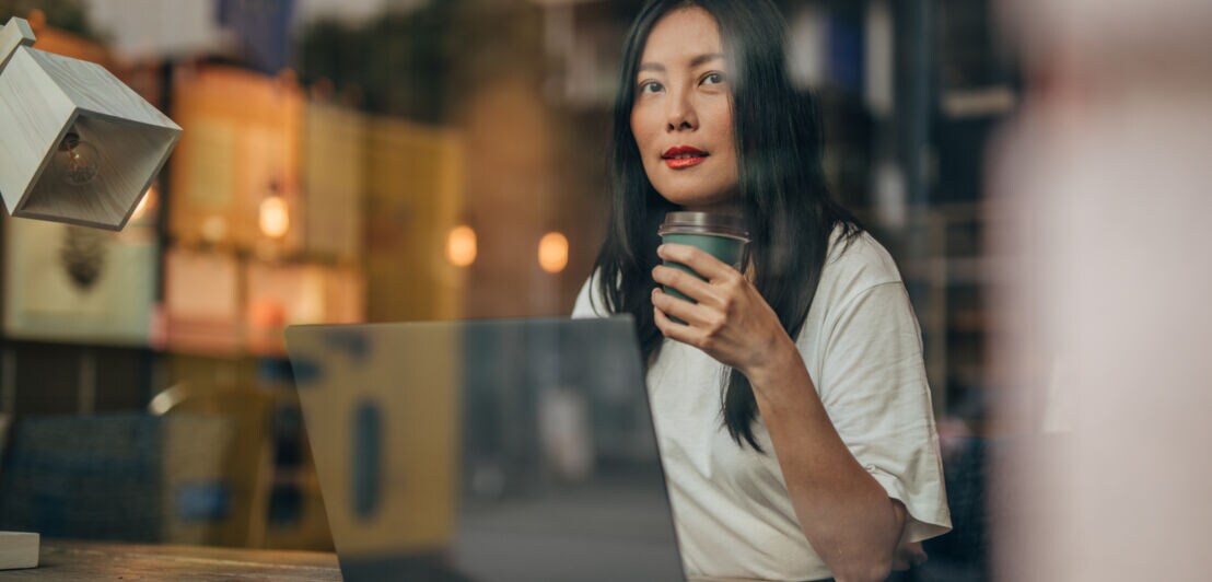 Eine Frau sitzt mit einem Coffee-to-go-Becher in einem Café am Laptop