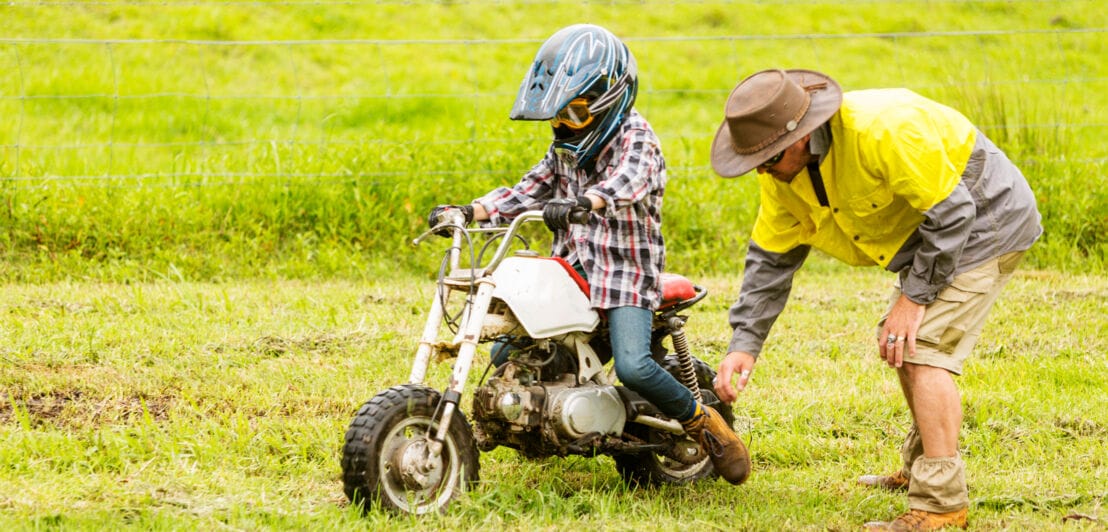 Kind auf Kindermotorrad auf einer feuchten Wiese, daneben ein gebückter Mann.