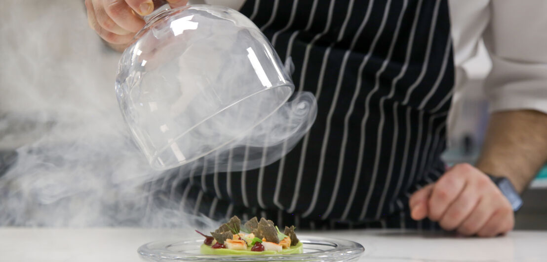 Die Hand eines Küchenchefs hebt eine Glasglocke von einem Teller mit einem exquisiten Gericht der Molekularküche