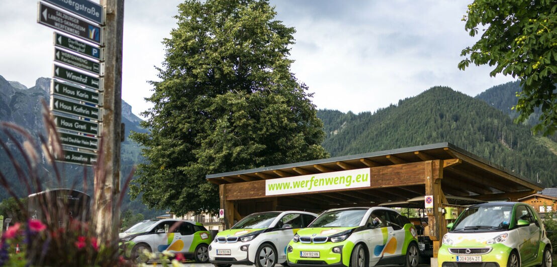 Mehrere E-Autos stehen unter einem Carport, während im Hintergrund begrünte Berge zu erkennen sind.
