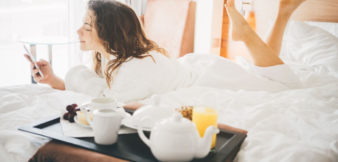 Eine Frau liegt im Bademandel auf einem Bett in ihrem Hotelzimmer. In der Hand hält sie ein Smartphone, neben ihr steht ein Tablett mit Frühstück.  