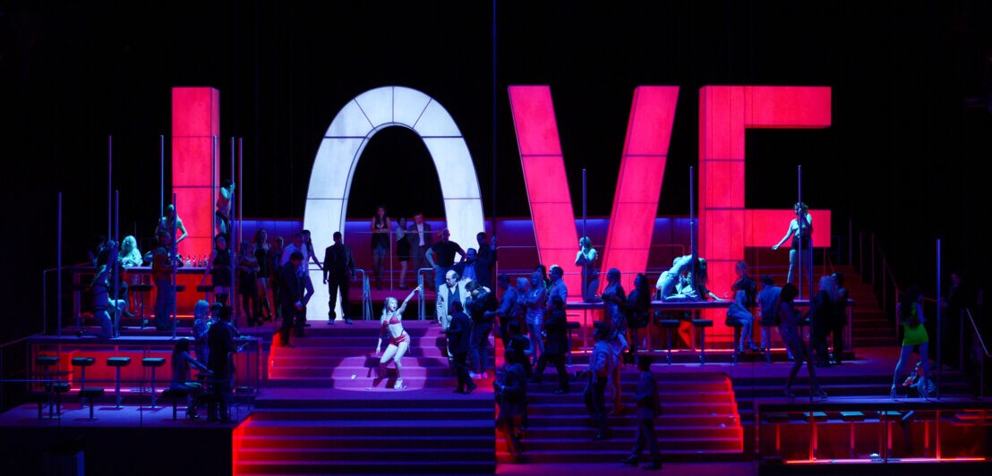 Darsteller:innen auf einer dunkel gehaltenen Theaterbühne vor einer rot-weißen Lichtinstallation aus vier Buchstaben, die das Wort Love bilden.