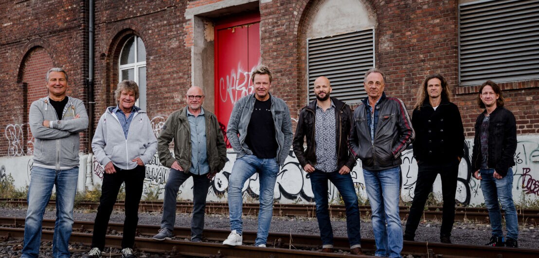 Die acht Mitglieder der Kölner Kultband Bläck Fööss stehen auf einem stillgelegten Bahngleis vor einem Backsteingebäude.