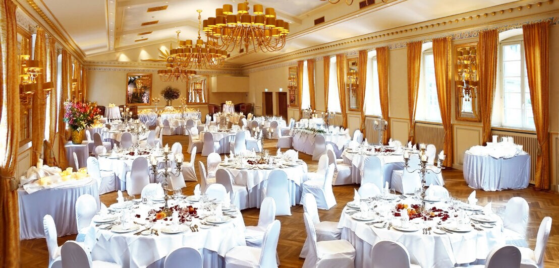 Stilvoll eingedeckte runde Tische mit weißen Stühlen in einem großen Festsaal mit Fischgrätenparkett.