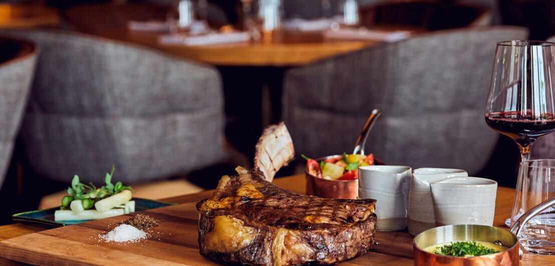 Ein gegrilltes Stück Fleisch liegt auf einem Holzbrett, im Hintergrund ist Restaurantambiente zu erkennen.