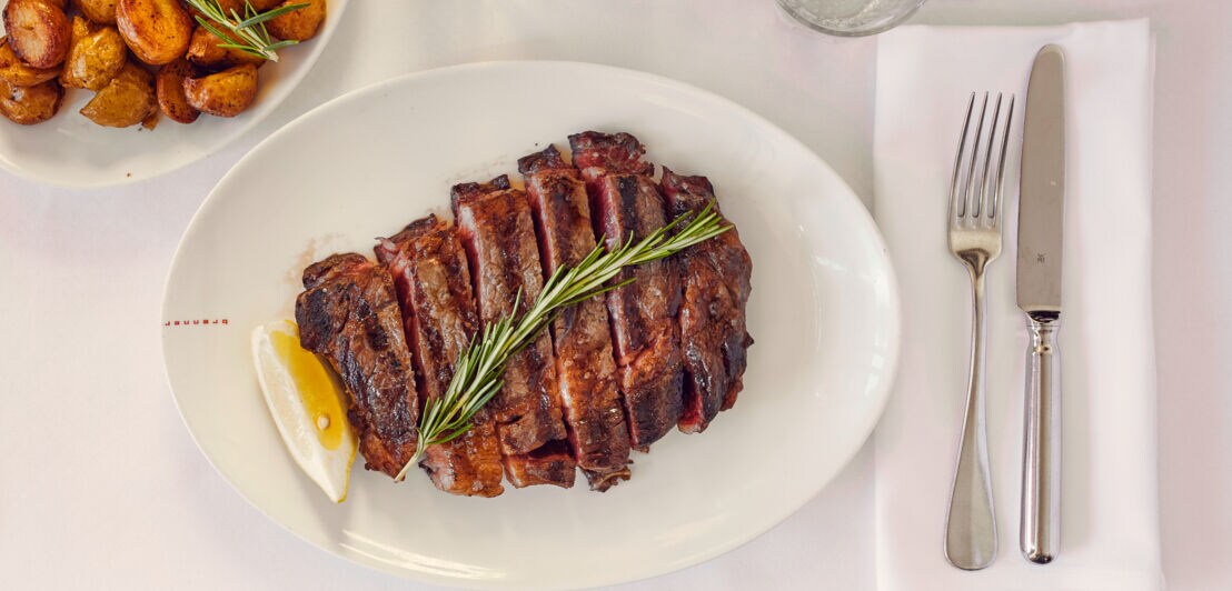 Ein geschnittenes Stück Fleisch liegt auf einem weißen Teller, daneben befindet Besteck sowie eine Schüssel mit Kartoffeln.