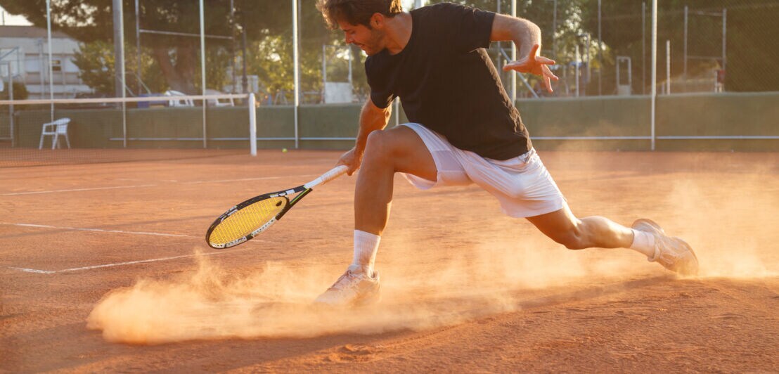 Eine Person mit Tennisschläger in der Hand bremst auf einem roten Schotterplatz.