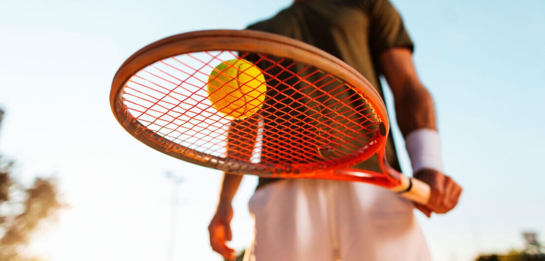 Eine sportlich gekleidete Person in der Froschperspektive fotografiert, die einen Tennisschläger über die Kamera hält, auf dem ein Tennisball liegt.