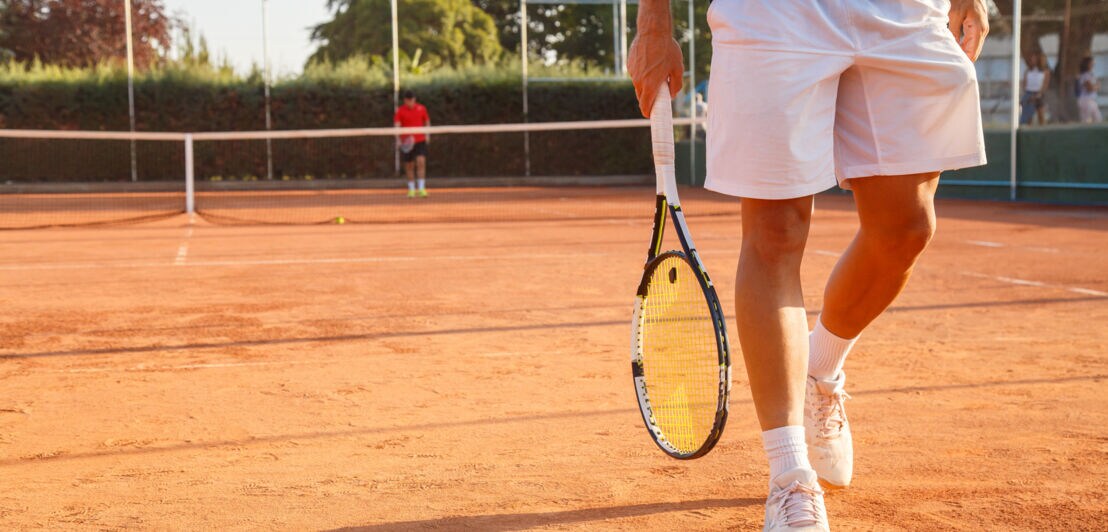 Eine Person geht über einen Tennisplatz und hat dabei einen Tennisschläger in der Hand.