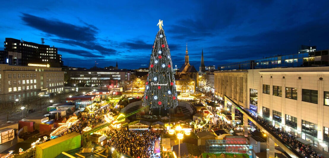 Blick von oben auf einen Weihnachtsmarkt in dessen Mitte ein riesiger beleuchteter Weihnachtsbaum steht