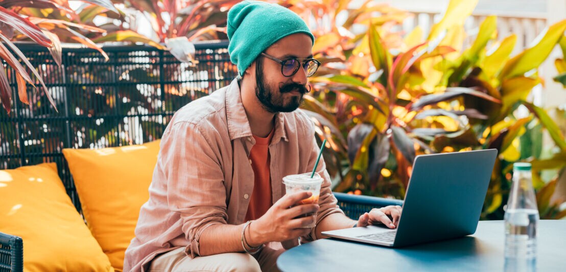 Ein junger Mann mit Bart und Mütze sitzt im Freien am Laptop, in der Hand hält er ein Getränk.