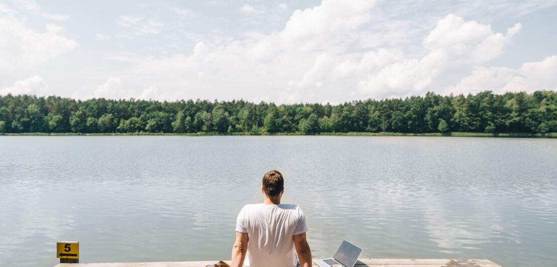 Ein Mann sitzt auf einem Steg am See und schaut aufs Wasser, neben sich ein Laptop.