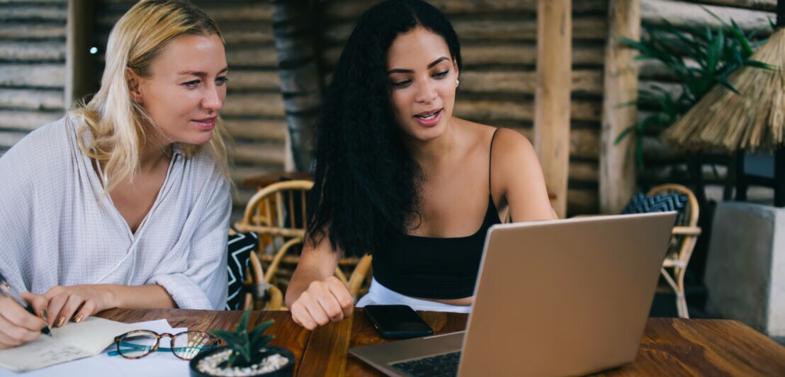 Zwei Frauen in Sommerkleidung sitzen an einem Holztisch vor einem Laptop.