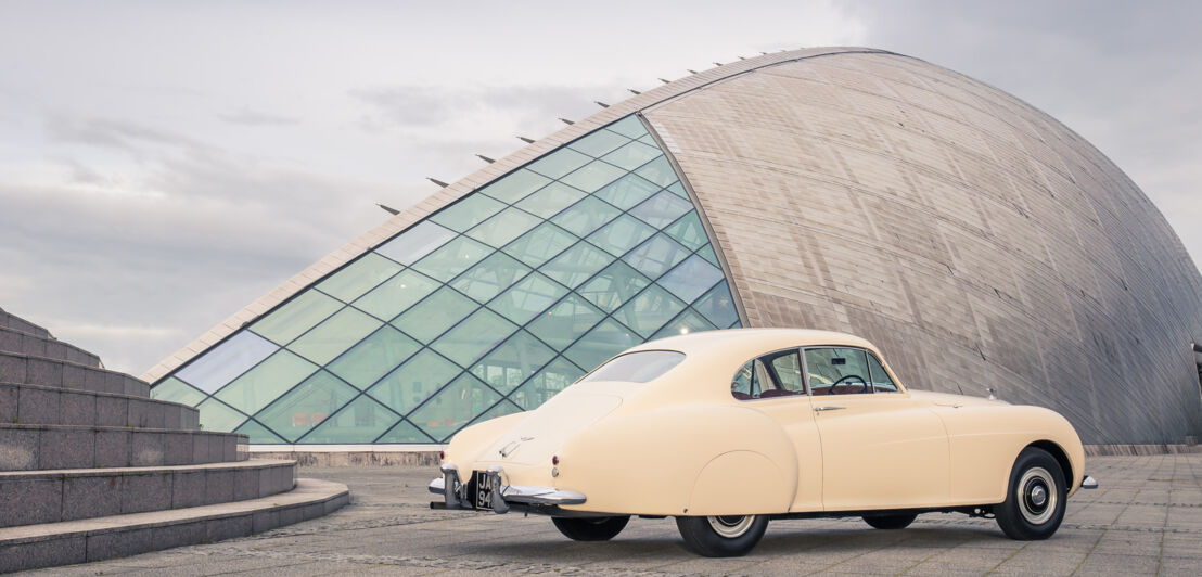 Ein cremefarbenes Auto von Bentley vor futuristischer Architektur