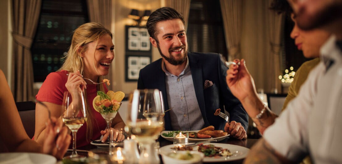 Eine blonde Frau und ein dunkelhaariger Mann bei einem Restaurantbesuch mit Freunden, vor ihnen ein gedeckter Tisch