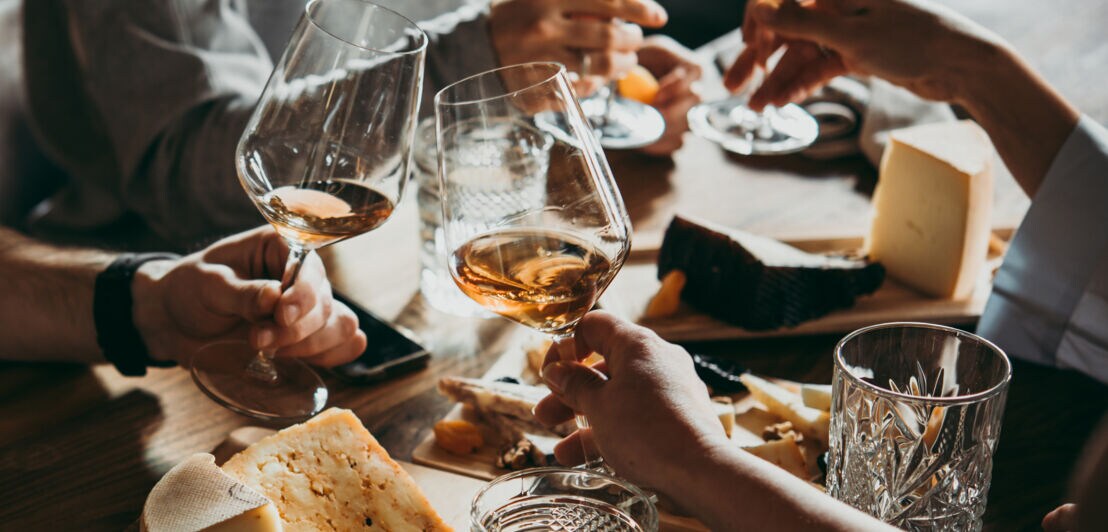 Mit Wein und Käse gedeckter Tisch, an dem mehrere Personen sitzen, die mit Gläsern anstoßen.
