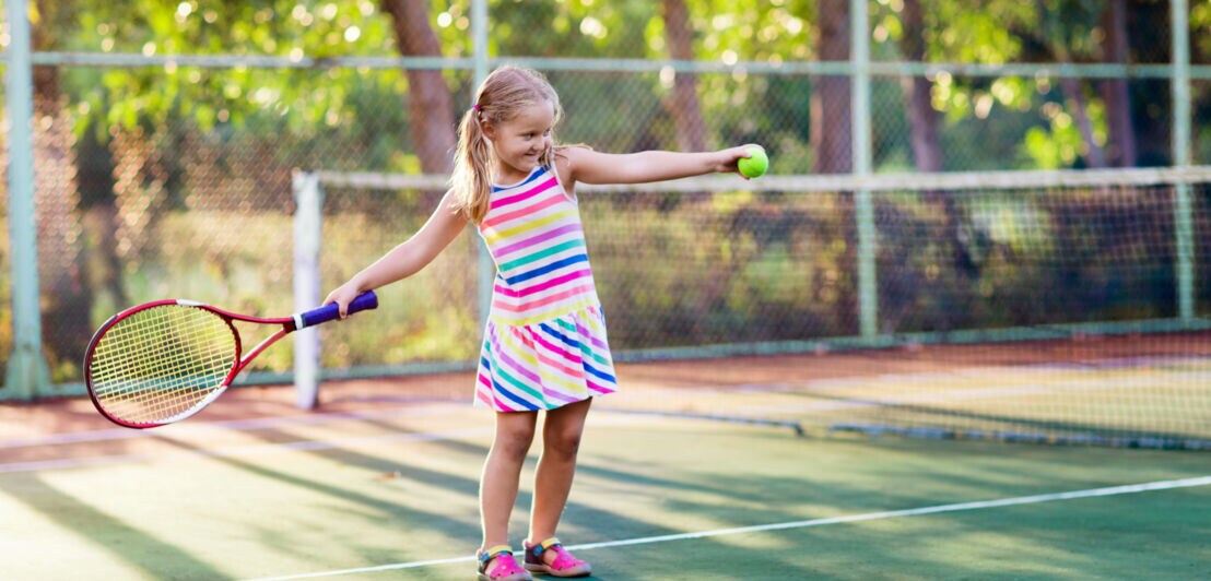 Ein kleines Kind im bunt gestreiften Kleid steht auf einem Tennisplatz und hält in der einen Hand einen Tennisschläger und in der anderen einen Tennisball.