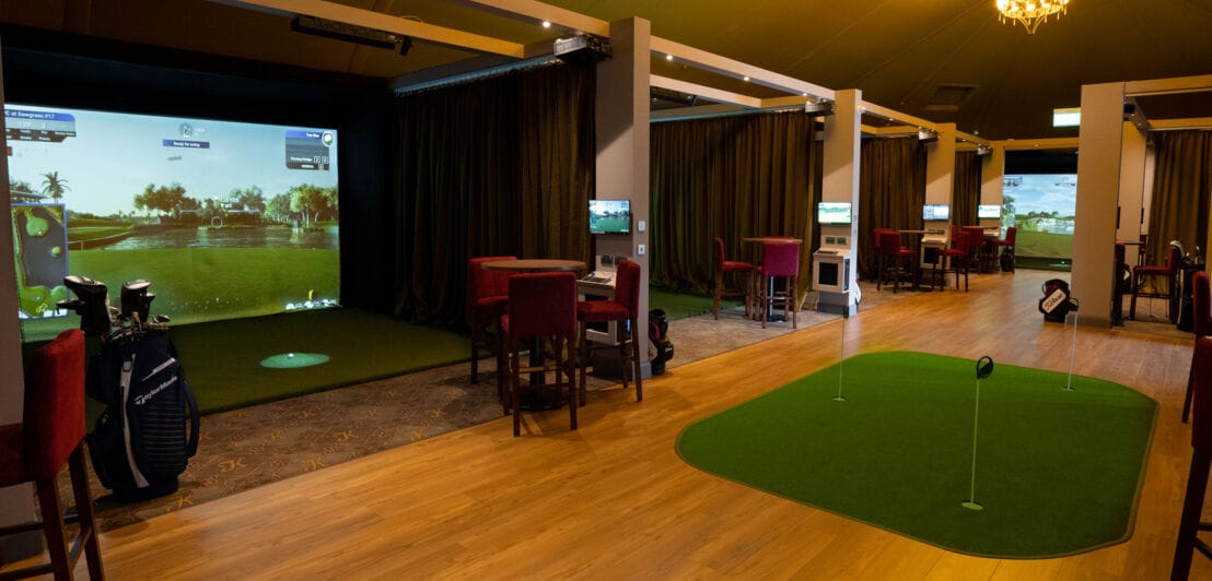 Ein großer Raum mit Holzboden, in dem mehrere Kabinen nebeneinander stehen, in denen ein digitaler Golfplatz gezeigt wird.