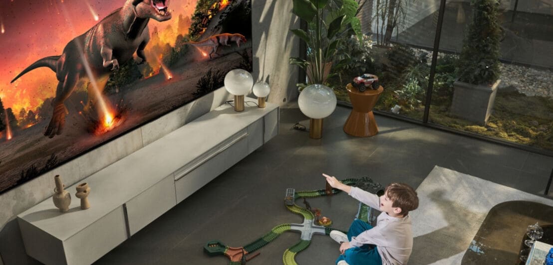Ein Junge sitzt in einem Wohnzimmer und zeigt auf einen großen Fernseher, auf dem ein Dinosaurier zu sehen ist