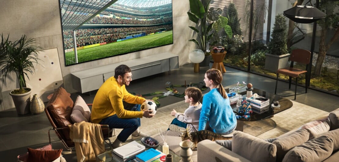 Eine Familie sitzt in einem Wohnzimmer vor einem großen Fernseher, auf dem ein Fußballfeld zu sehen ist, der Mann lächelt das Kind und die Frau an und hält einen Fußball in den Händen