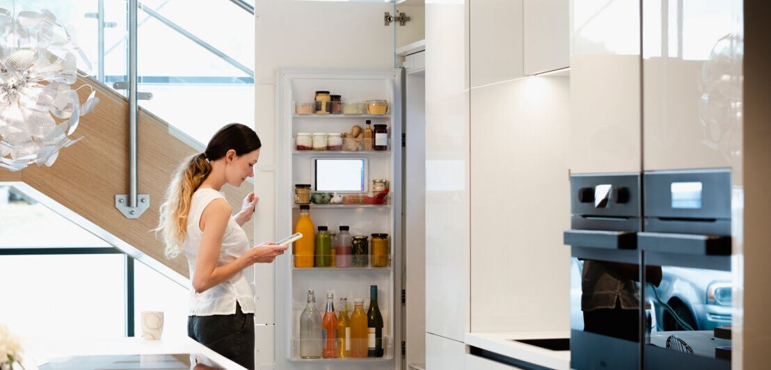 Seitenansicht einer Person, die vor einem geöffneten Kühlschrank steht und auf ihr Smartphone schaut