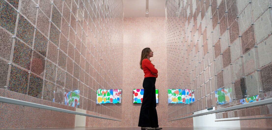 Eine Frau betrachtet gepunktete Kunstwerke hinter einer hängenden Plexiglaswand in einer Galerie, im Hintergrund digitale Kunstwerke auf Monitoren