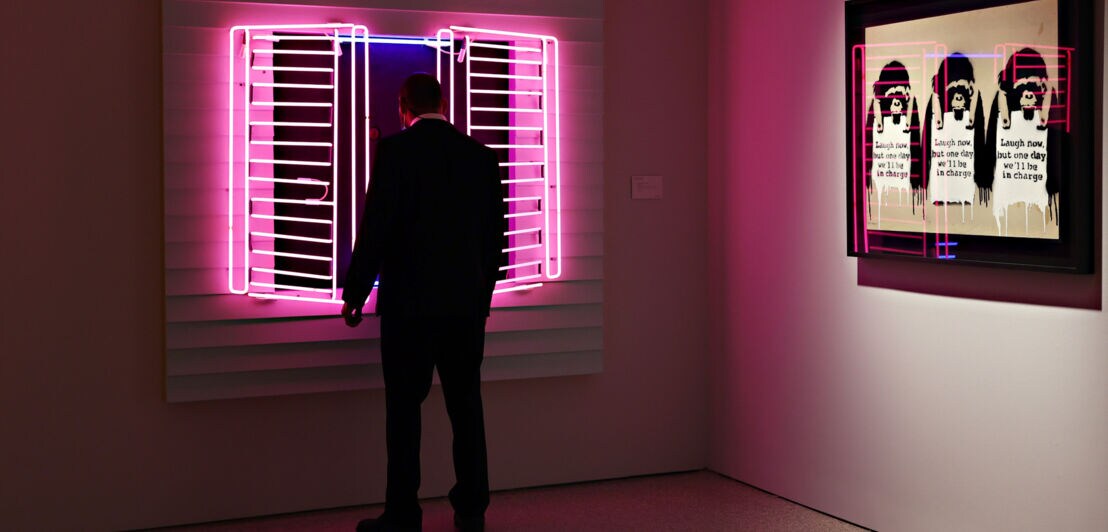 Eine Person schaut sich ein Kunstwerk in der Form eines Fensters aus Neonröhren an der Wand eines Ausstellungsraums an