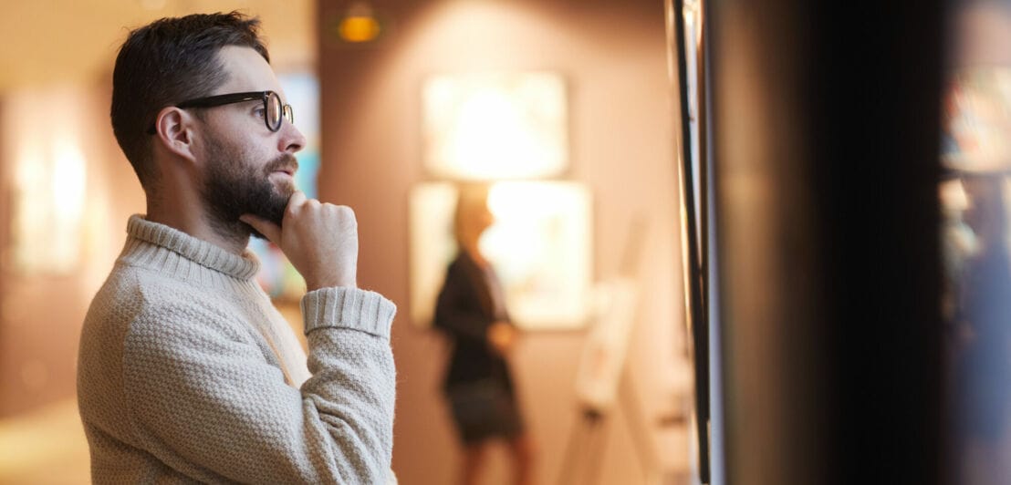 Ein Mann mit Brille und Rollkragenpullover betrachtet interessiert ein Kunstwerk in einer Galerie