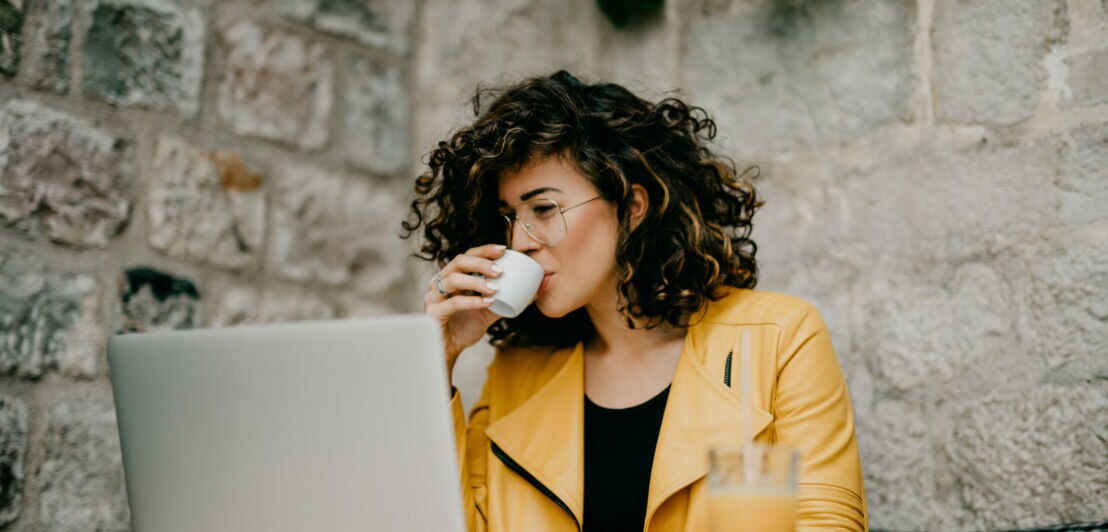 Eine Frau in gelber Jacke mit lockigen schwarzen Haaren und Brille sitzt im Café am Laptop und trinkt einen Espresso
