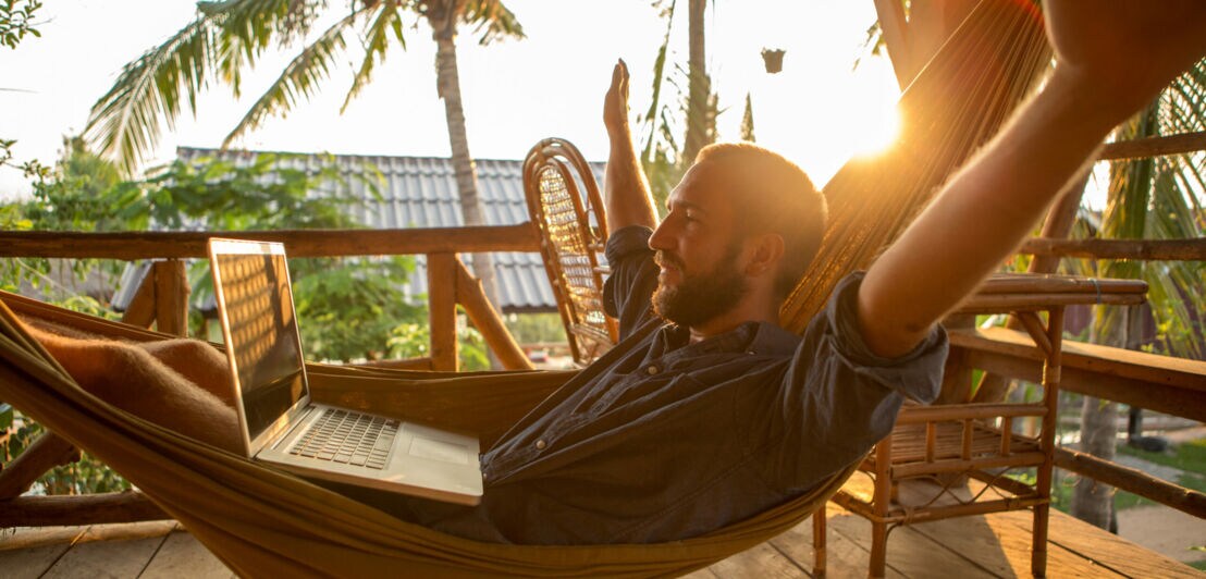 Ein Mann mit Bart liegt in der Hängematte in einem tropischen Land und schaut auf den Laptop auf seinem Schoß, die Arme streckt er aus