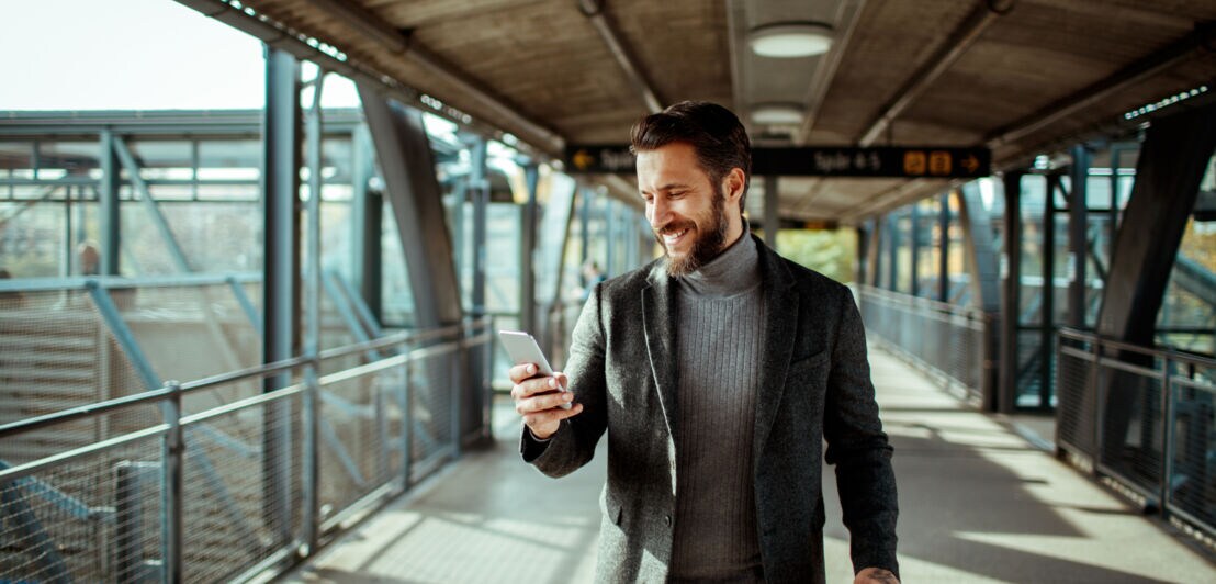 Ein Mann mit Bart und im Sakko schaut am Flughafen auf sein Handy