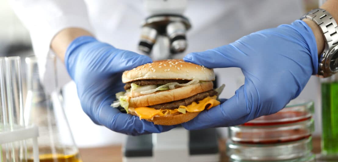 Eine Person im weißen Kittel mit blauen Gummihandschuhen hält einen Burger vor einem Mikroskop fest