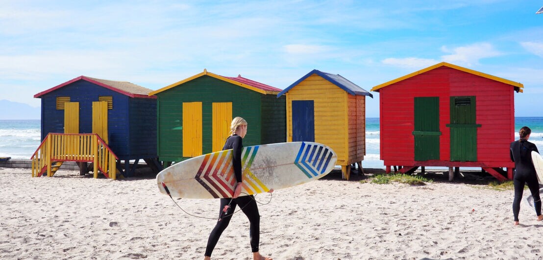 Surferinnen vor bunten Umkleidekabinen aus Holz an einem Strand bei Kapstadt