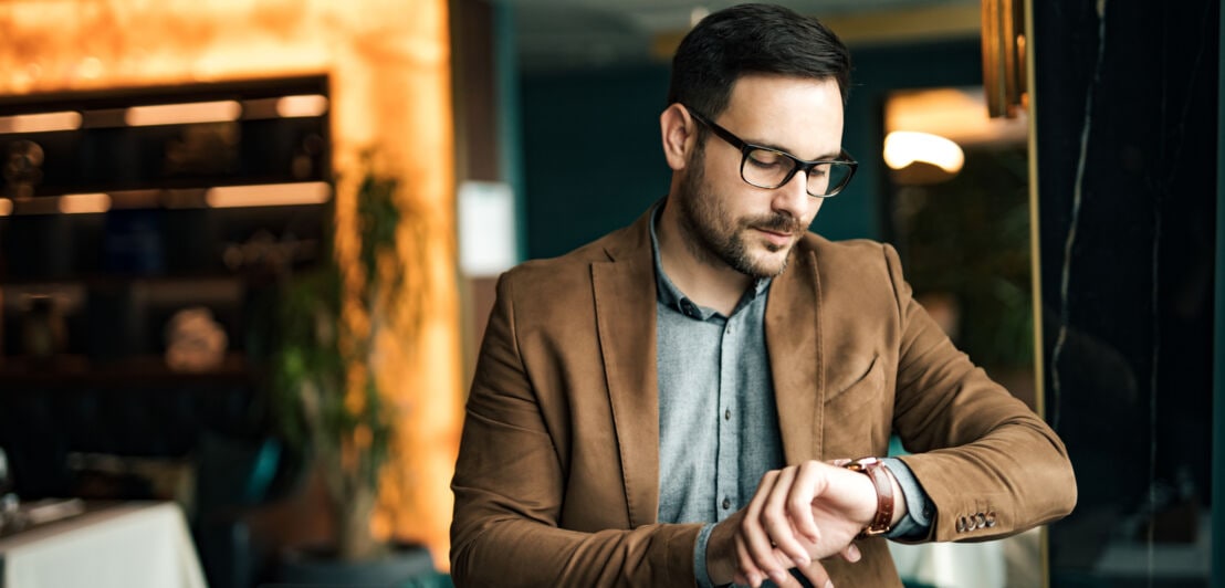 Ein Mann im Jacket, mit Brille und Bart schaut auf seine Armbanduhr