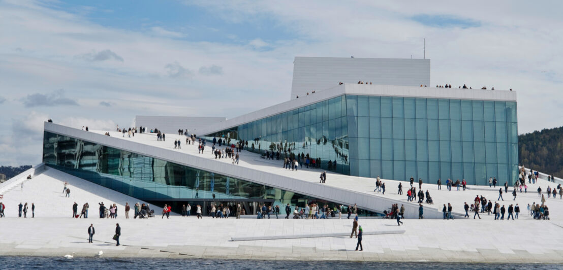 Personen laufen auf einem weißen, futuristischen Gebäude mit Glasfassade am Wasser