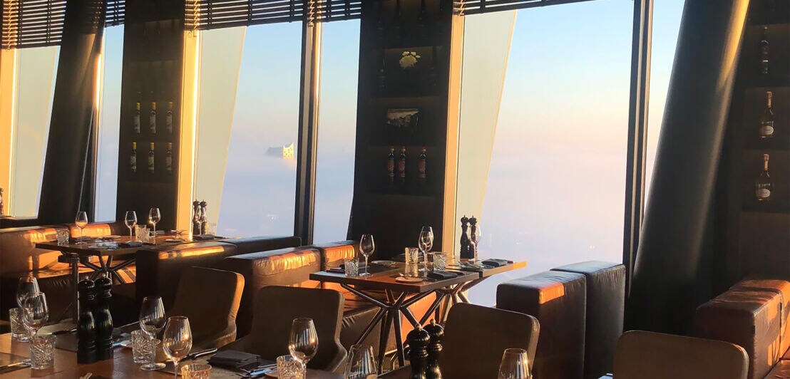 Innenaufnahme eines gehobenen Restaurants mit gedeckten Tischen und Panoramafenstern mit Blick auf die wolkenverhangene Hamburger Elbphilharmonie