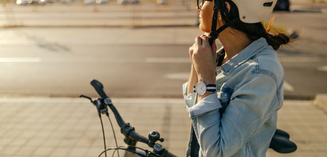 Eine Person steht mit ihrem Fahrrad an einer Straße und schließt gerade ihren Helmgurt