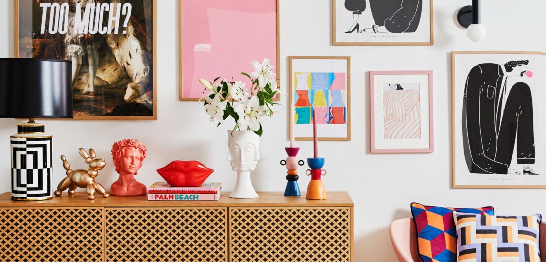 Eine gemusterte Holzkommode mit verspielten Accessoires obendrauf und ein rosa Stuhl mit gemusterten Kissen vor einer Wand mit gerahmten Bildern