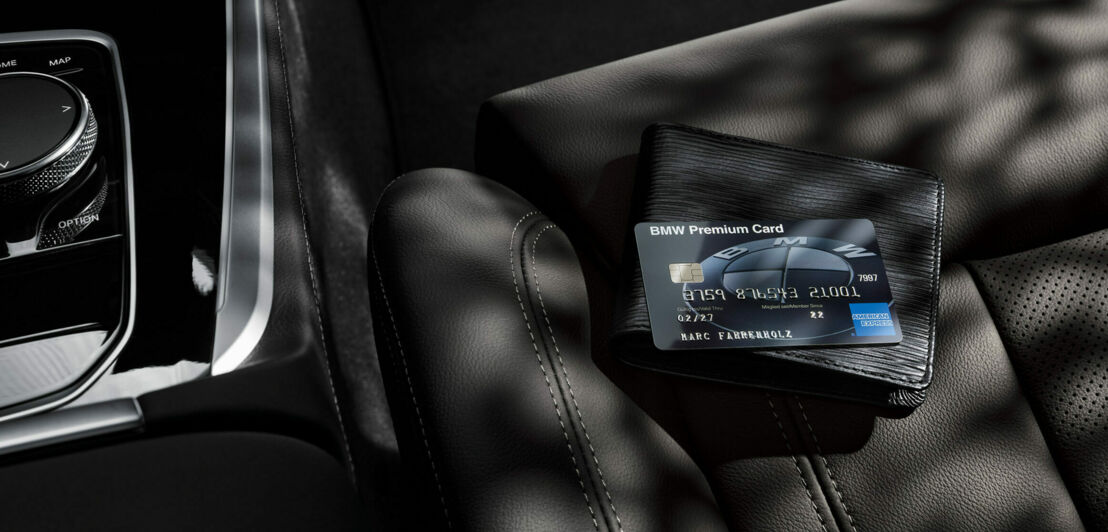 Eine schwarze BMW Premium Card von American Express liegt auf einem schwarzen Portemonnaie auf einem schwarzen Autositz aus Leder