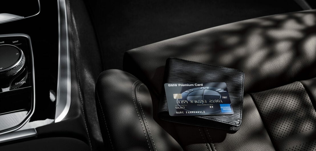 Eine schwarze BMW Premium Card von American Express liegt auf einem schwarzen Portemonnaie auf einem schwarzen Autositz aus Leder