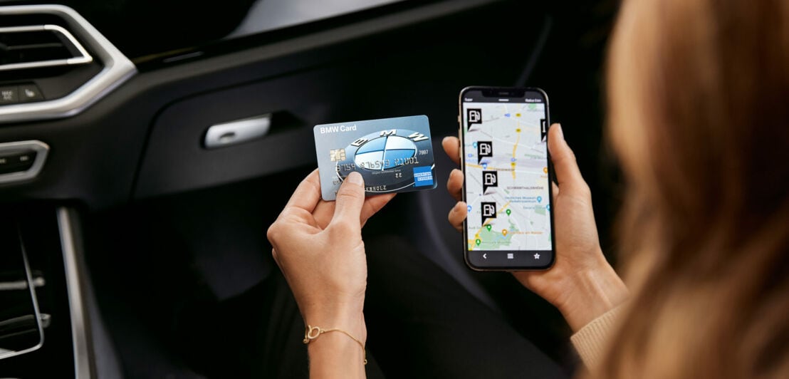 Eine Frau in einem Auto hält eine BMW Card von American Express neben ein Smartphone mit einer geöffneter App, die Tankstellen anzeigt