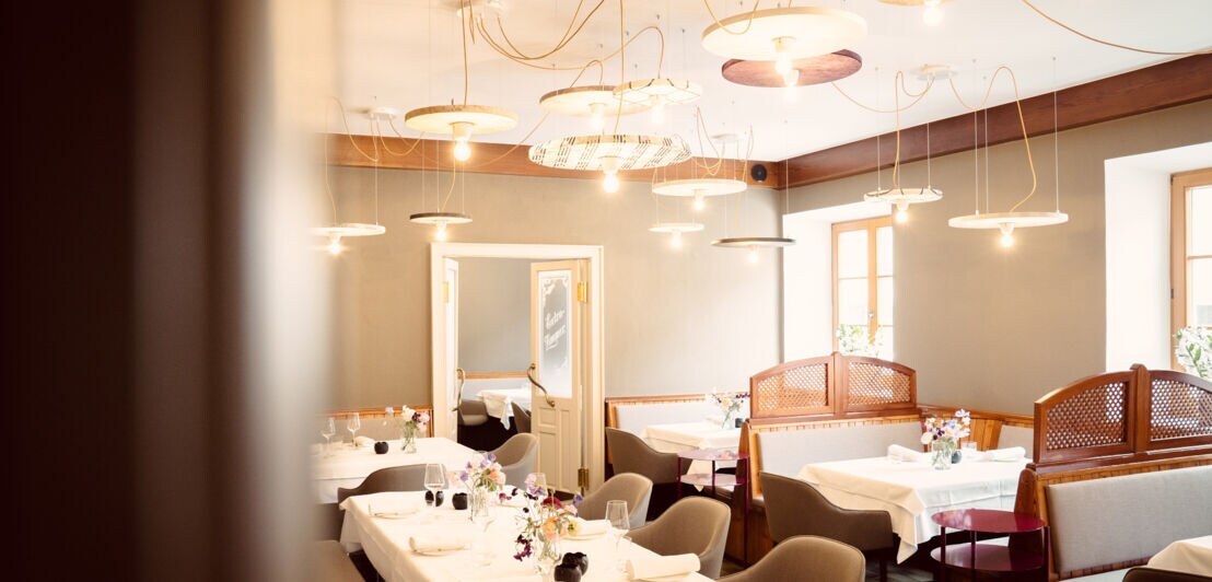 Der Speiseraum eines modern gestalteten Wirtshauses mit weiß eingedeckten Tischen, gepolsterten Stühlen und vielen runden Deckenlampen
