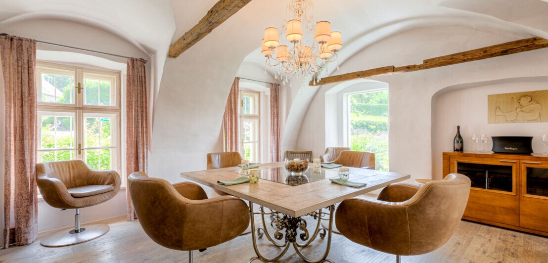 Ein heller Raum mit Fenstern, in dessen Mitte ein viereckiger, eingedeckter Tisch mit braunen Ledersesseln steht