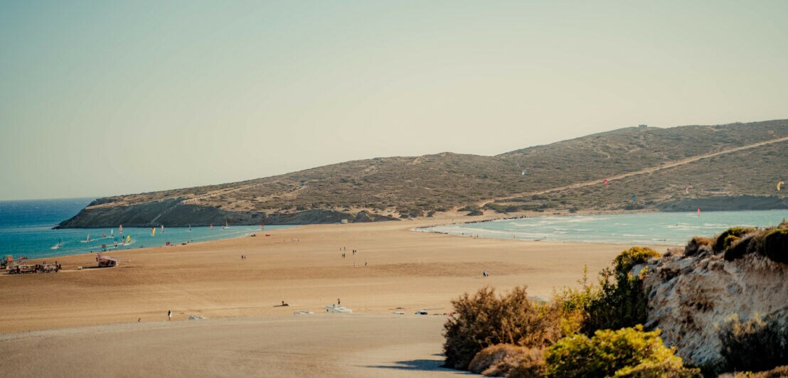 Ein sonniger Tag am Strand von Prasonisi. Die schmale Sandbank führt zur kleinen hügeligen Halbinsel und auf dem Wasser sowie in der Luft tummeln sich bunte Surfer:innen und Kitesurfer:innen.