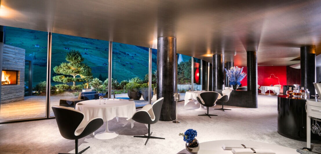 Ein großer Raum mit stylish-elegantem Interieur, weiß gedeckten Tischen und einer großen Glaswand, im Hintergrund ein Kamin