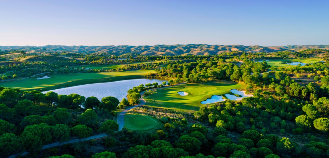 Ein Golfplatz mit vielen Bäumen und Wasserhindernissen von oben, im Hintergrund Berge