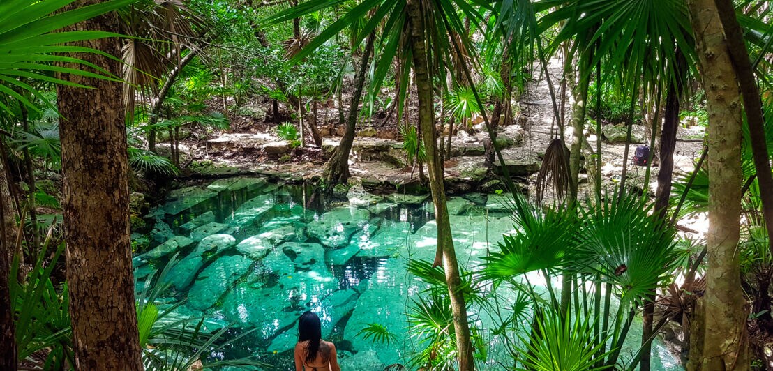 Eine Frau im schwarzen Bikini sitzt im Dschungel am Wasser, umgeben von Palmen